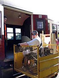 Photo of Bill sitting in his chair, in a metal enclosure, being lifted up to a wide side entry of a train car.
