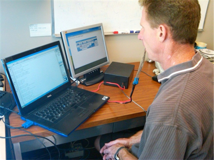 Title: A photo of Kurt Manal using the Mouth Mouse. - Description: The photo shows a side view of Dr. Manal with a flat cable, connected to a computer before him, protruding from his mouth.