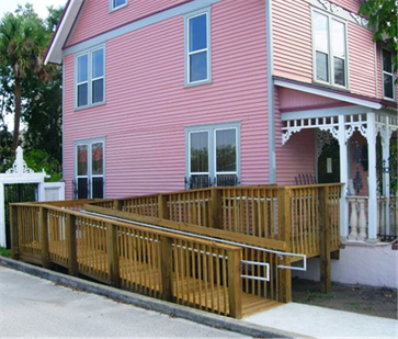 Title: A photo of a wooden exterior ramp at the side of a house. - Description: The photo shows a wooden ramp beside a pink house. The ramp rises from a cement walkway, is constructed of attractive wood slats and has metal handrails on both sides. The ramp has one switchback and rises several feet to the front porch.